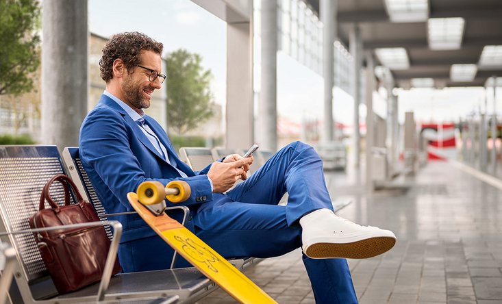 Ein sportlicher Business-Mann sitzt auf einer Bank am Bahnsteig und schaut lächelnd auf sein Handy.