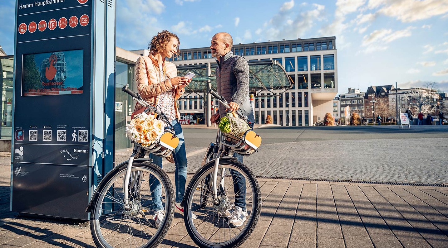 Eine Frau und ein Mann mit Leihrädern stehen an einer Stele einer Mobilstation.