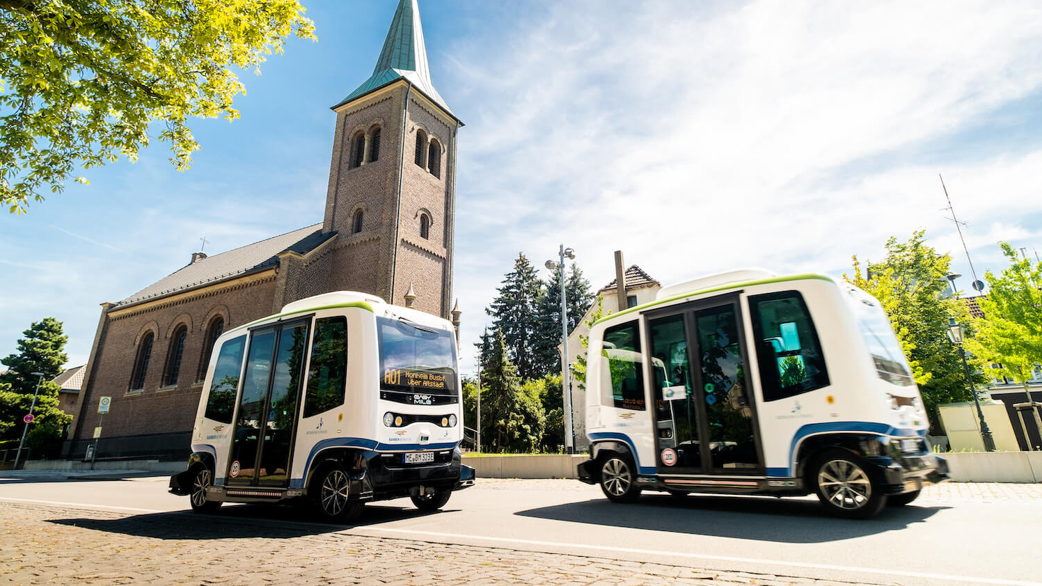 Zwei autonome Kleinbusse fahren auf Kopfsteinpflaster an einem sonnigen Tag, im Hintergrund ist eine Kirche zu sehen.