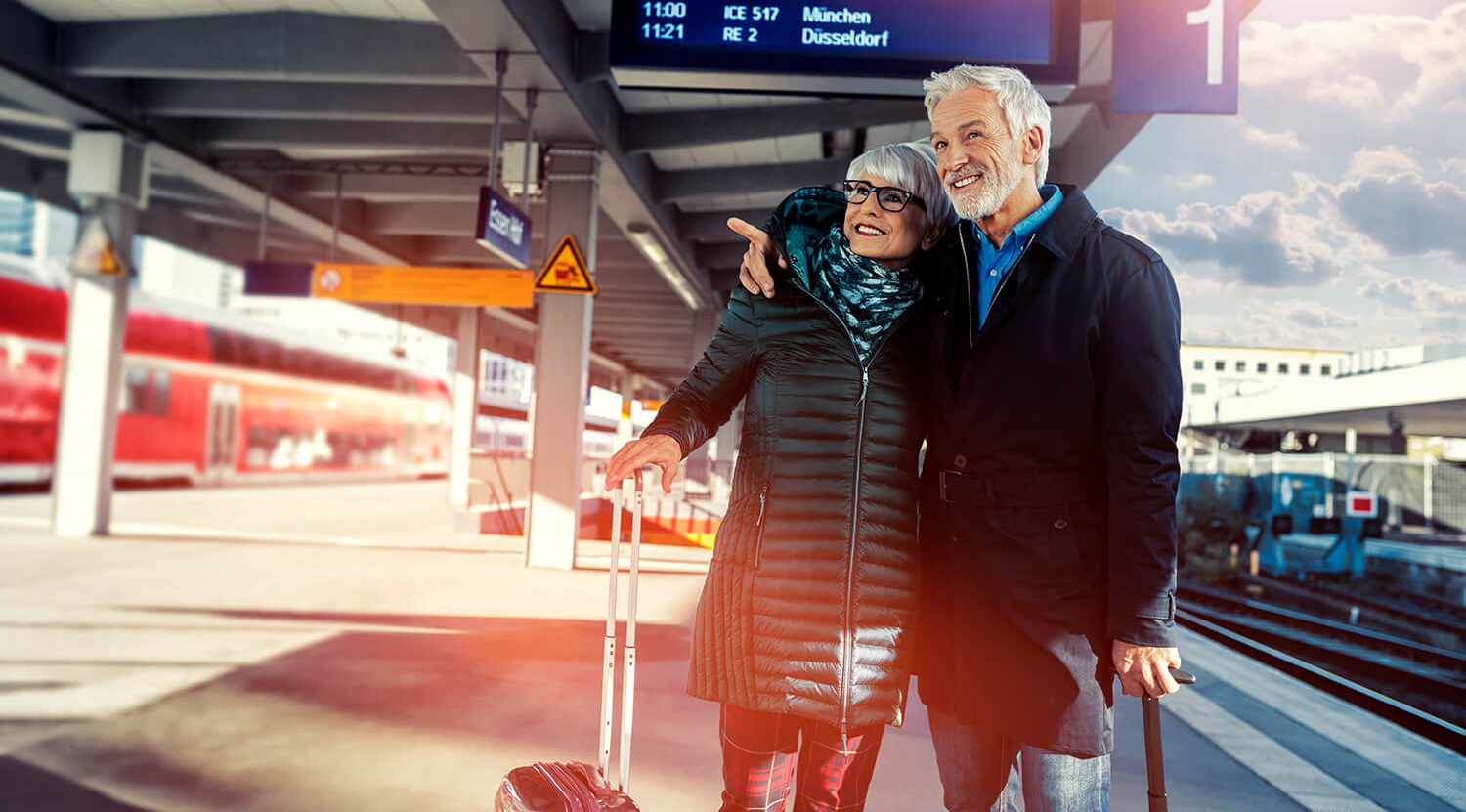 Ein älteres Ehepaar steht mit Koffern an einem bahnsteig und blickt die Gleise entlang.