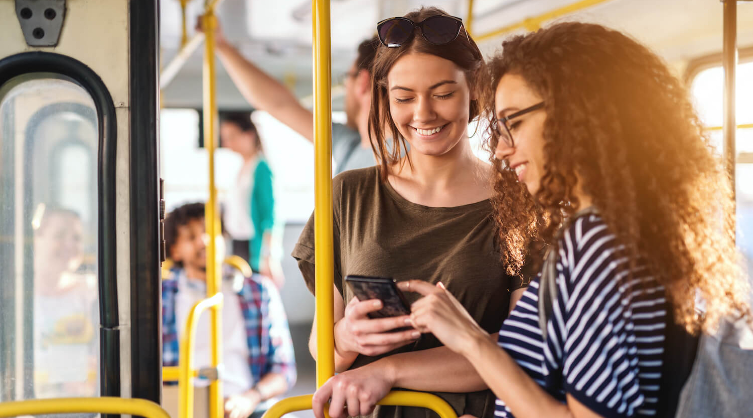 Zwei junge Frauen stehen gut gelaunt in einem Bus und schauen auf ein Smartphone.