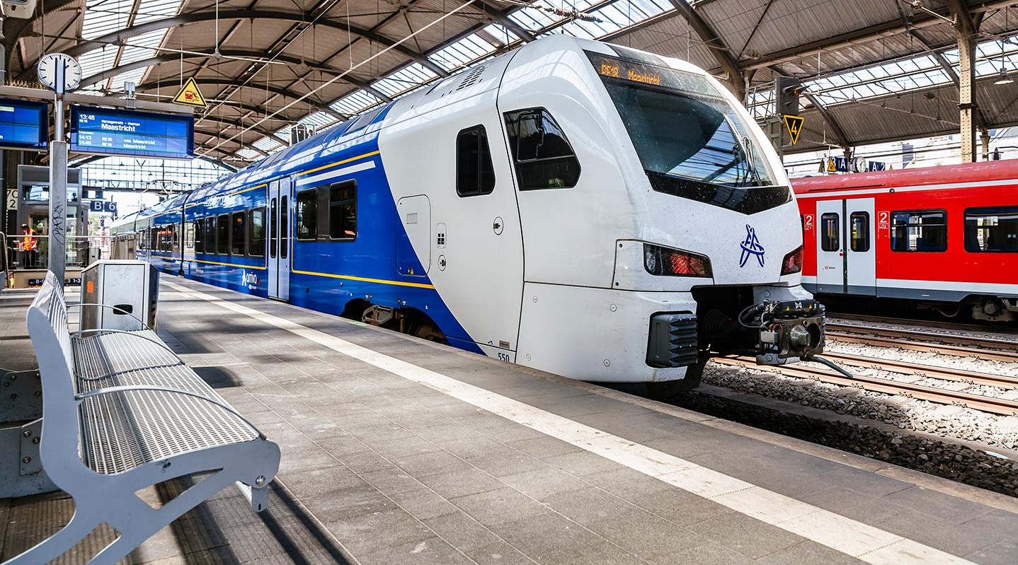 Ein blau-weißer Zug steht an einem überdachten Bahnhof am Gleis. Im Hintergrund ist ein roter Zug zu sehen.