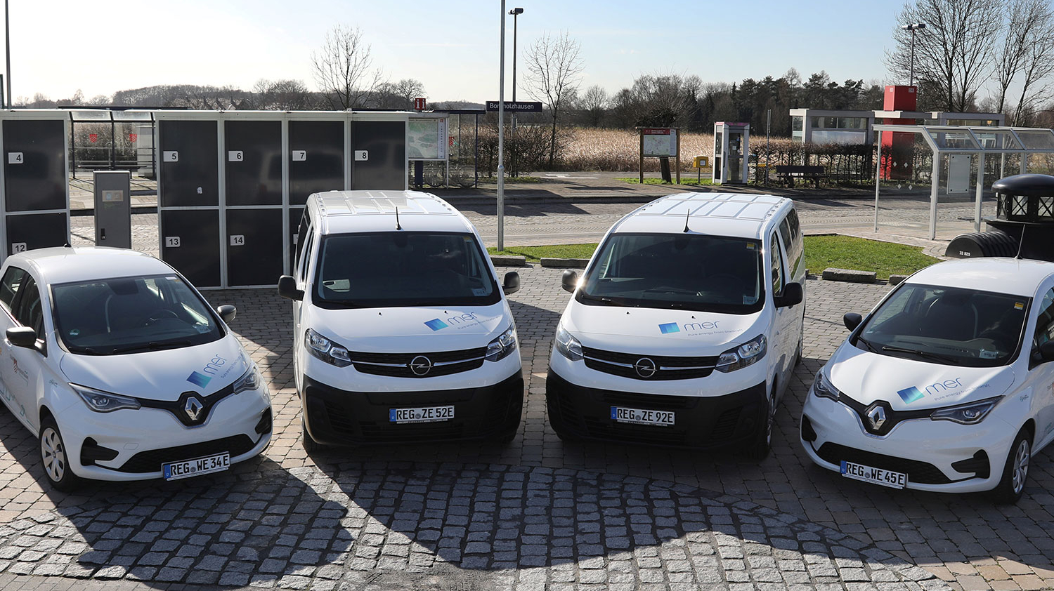 Vier E-Autos stehen auf einem Kopfsteinplatz vor den Gleisen des Bahnhofs Borgholzhausen.