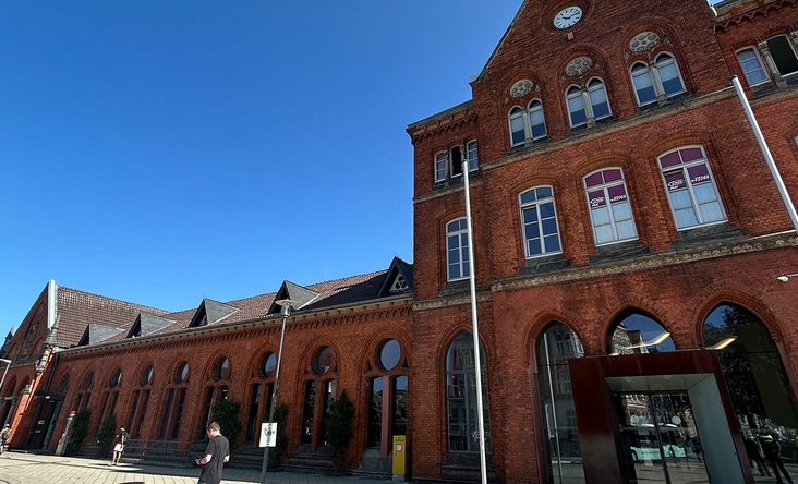 Historischer Backstein‑Bahnhof Detmold mit hohen Rundbogenfenstern und Uhrgiebel, sonniger Vorplatz unter blauem Himmel