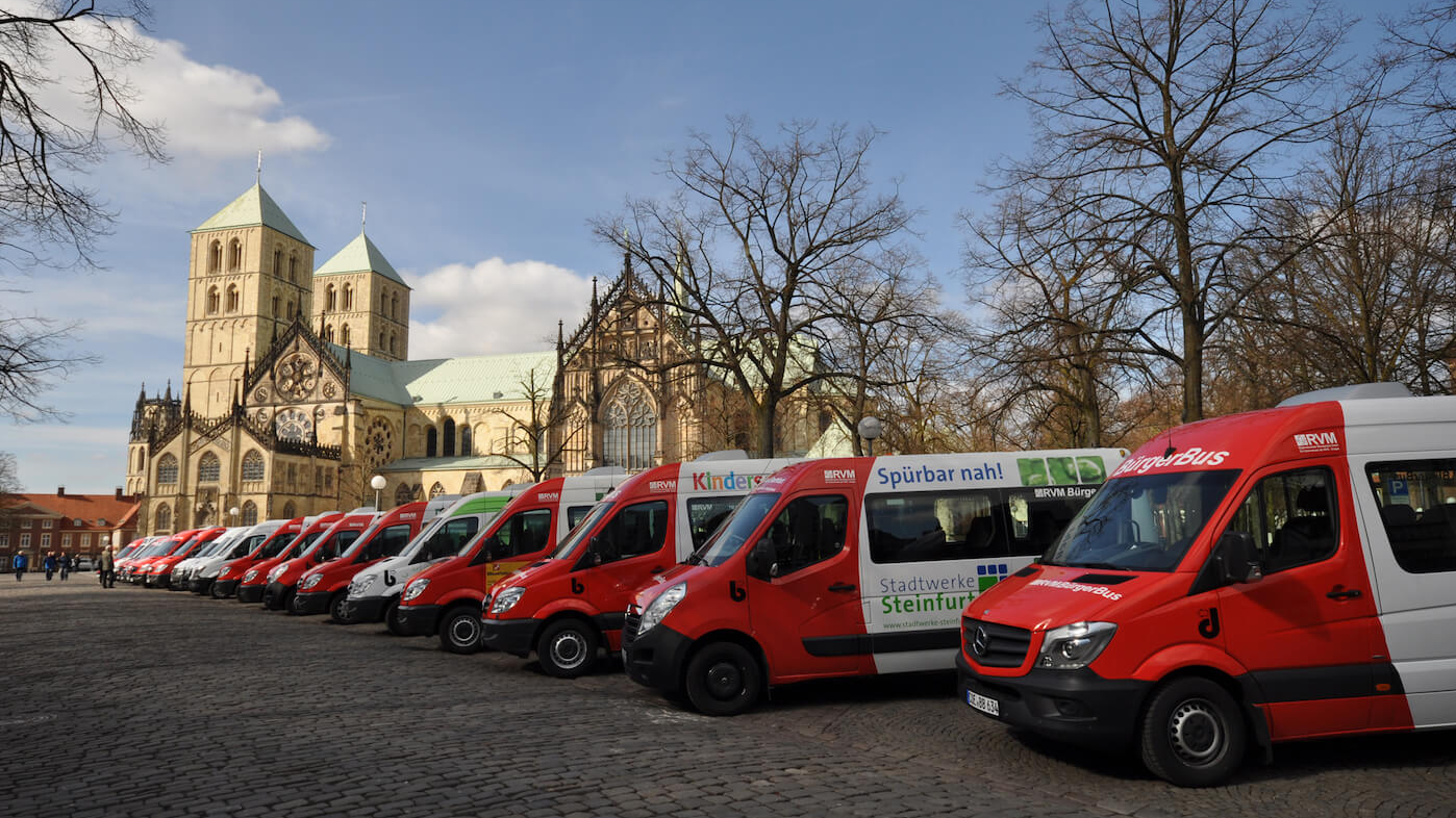Eine Reihe roter Kleinbusse steht nebeneinander auf einem Parkplatz, im Hintergrund sieht man ein altes Gebäude.