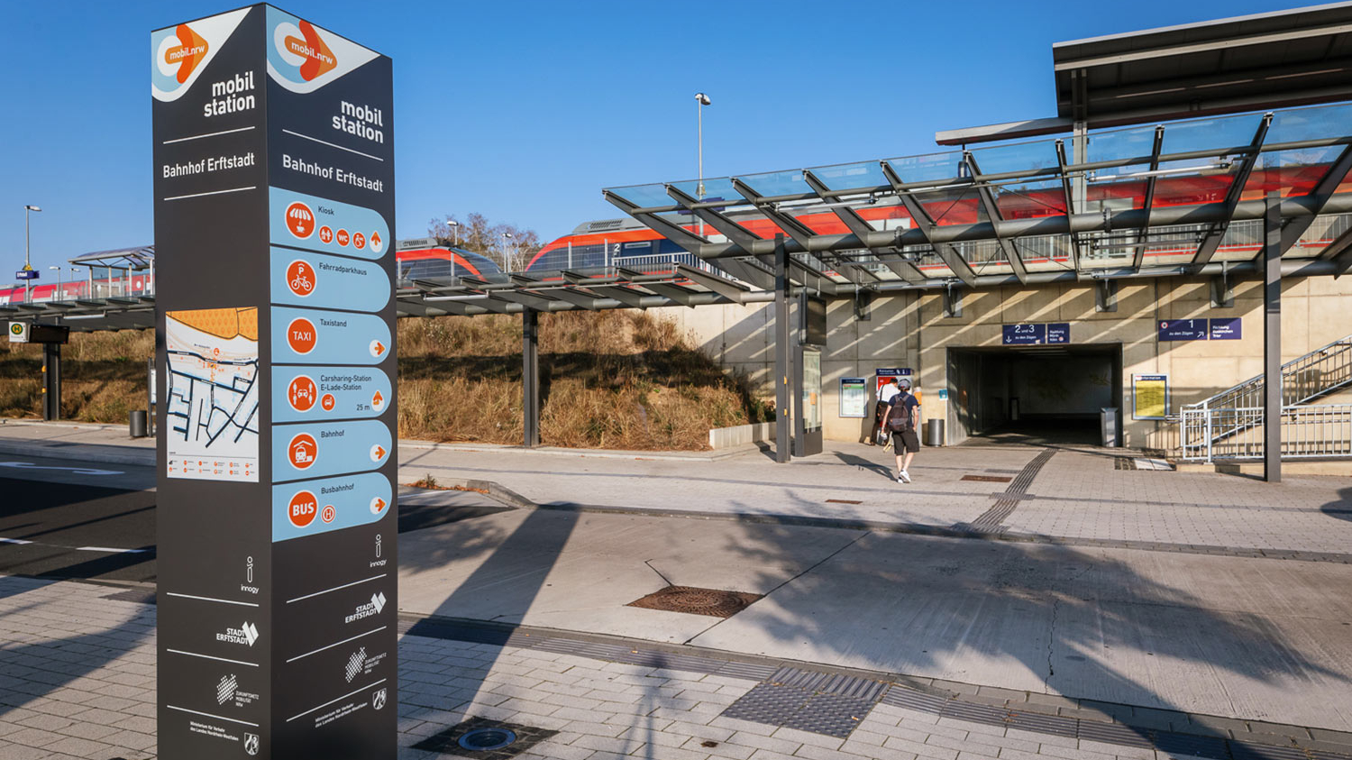 Die Säule der Mobilstation am Bahnhof Erftstadt. Im Hintergrund fährt ein Zug vorbei.