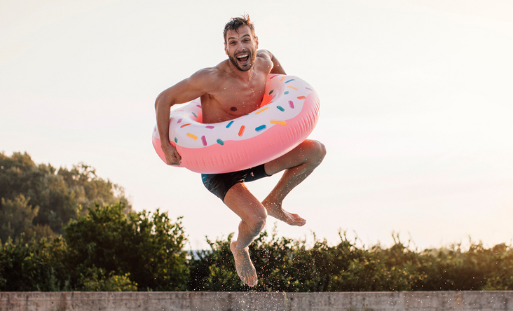 Ein Mann mit einem Schwimmring springt in einem Freibad fröhlich vom Beckenrand ins Wasser.