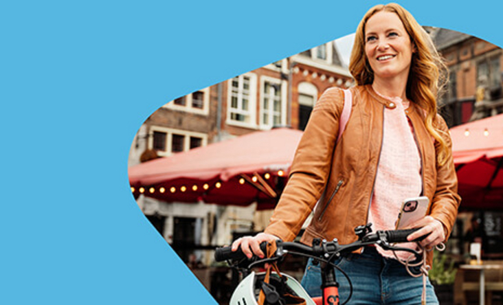 Zu sehen ist eine junge Frau mit blonden Haaren, welche ein Fahrrad durch eine Stadt schiebt. 