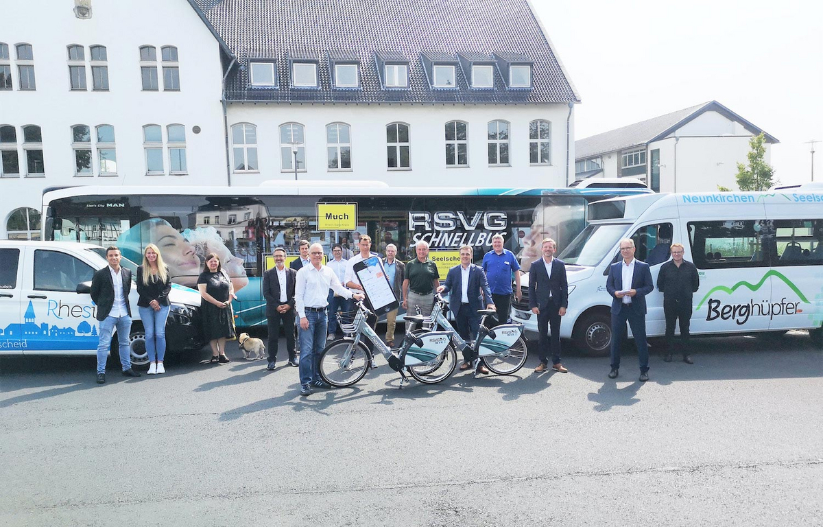 Ein Gruppenbild mit mehreren Menschen, die vor verschiedenen Kleinbussen und an Leihrädern stehen.