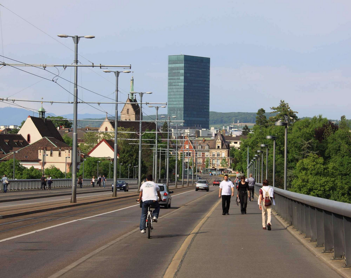 Fußgänger*innen und Radfahrer*innen bewegen sich über eine Brücke. Im Hintergrund sieht man Hochhäuser und Häuser in Basel.