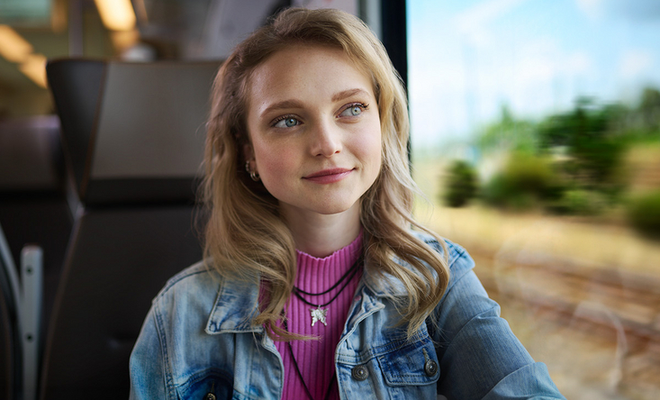 Eine junge Frau sitzt in einem Zug am Fenster und schaut lächelnd hinaus.