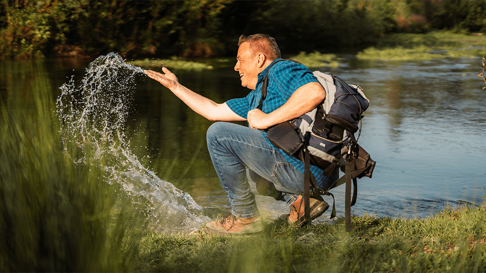 Zu sehen ist ein Mann, welcher am Rande eines GEwässers steht und mit seiner Hand das Wasser in die Luft befördert. 