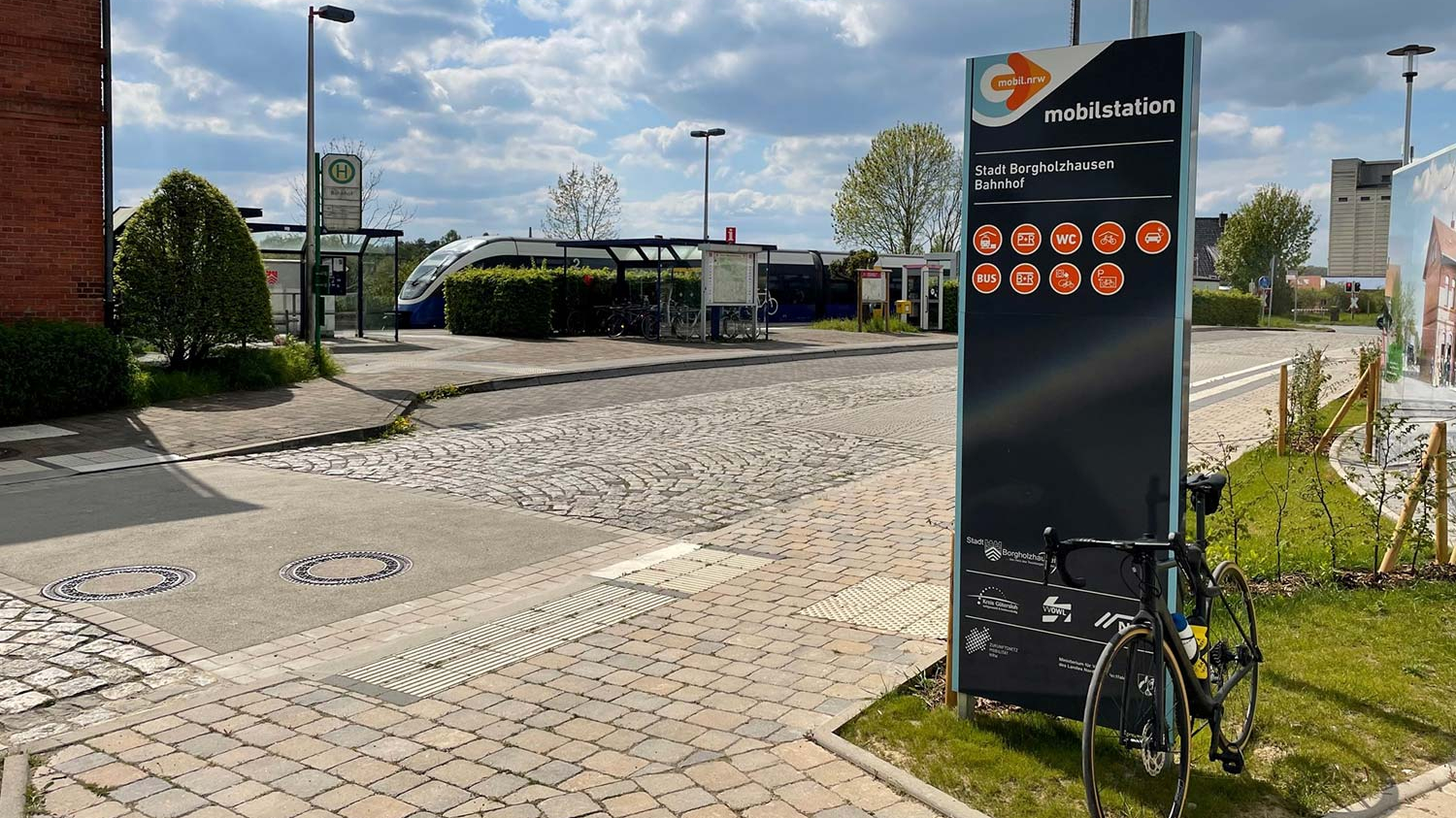 Ein Fahrrad lehnt an der Stele der Mobilstation Borgholzhausen, während im Hintergrund ein Zug fährt.