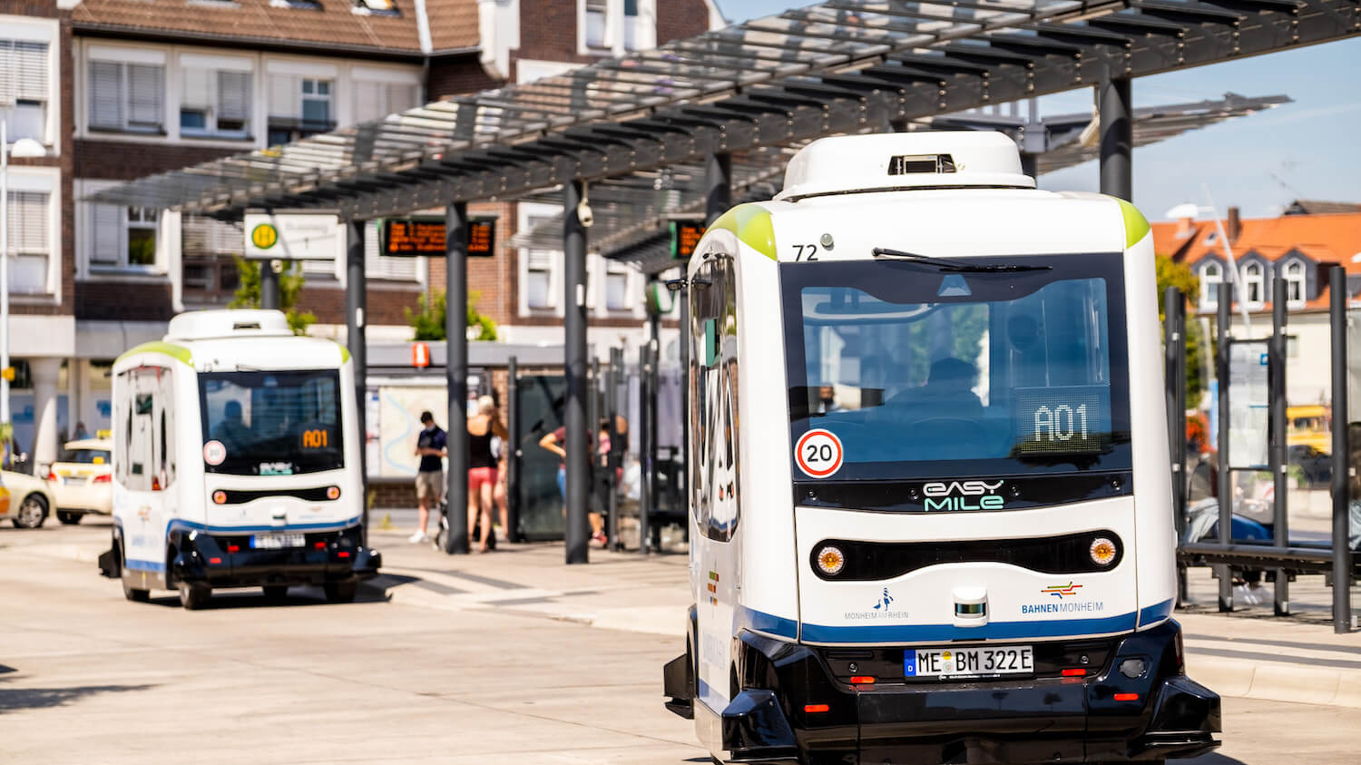 Zwei hintereinander fahrende autonome Kleinbusse an einem Busbahnhof.