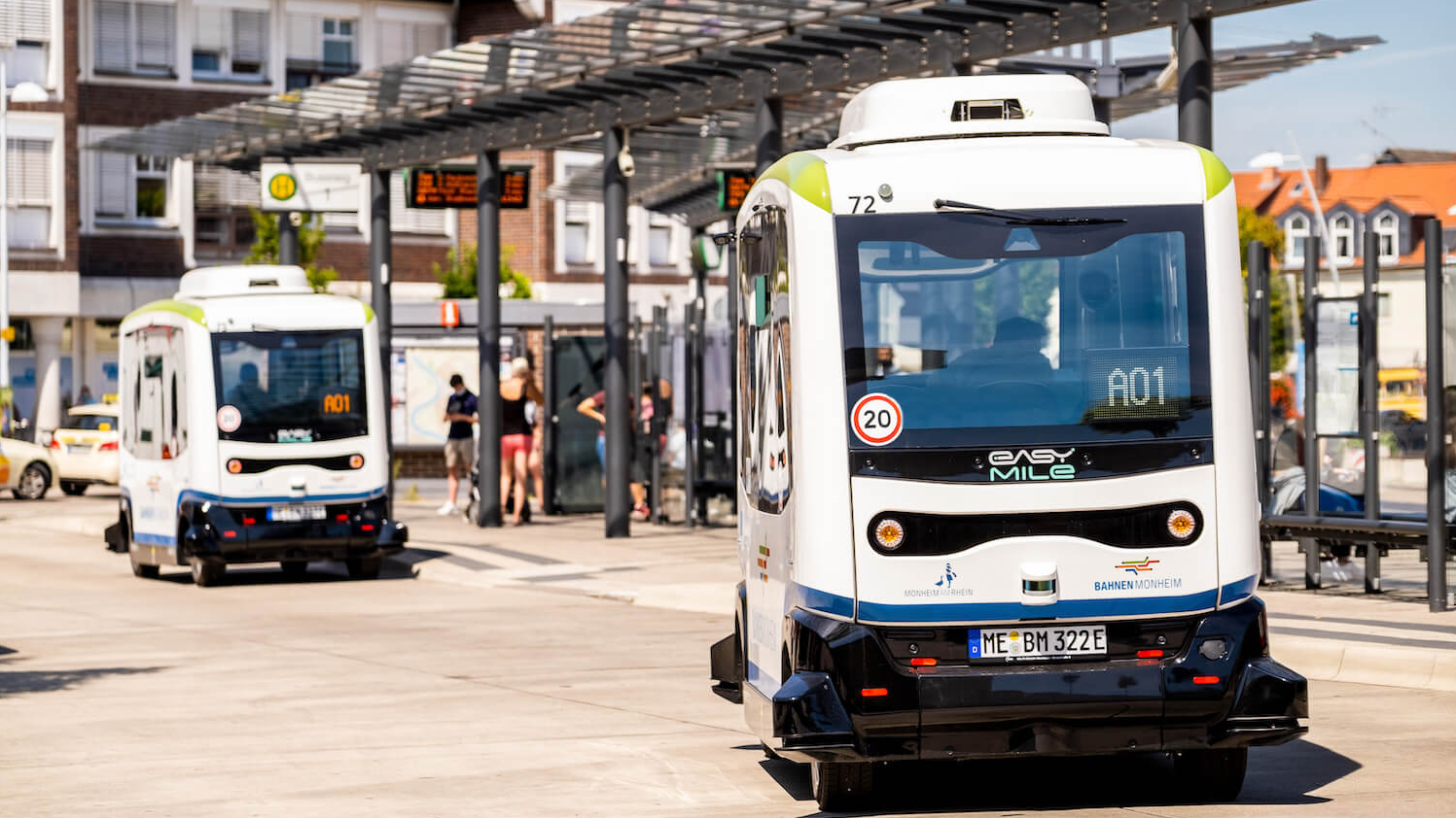 Zwei hintereinander fahrende autonome Kleinbusse an einem Busbahnhof.