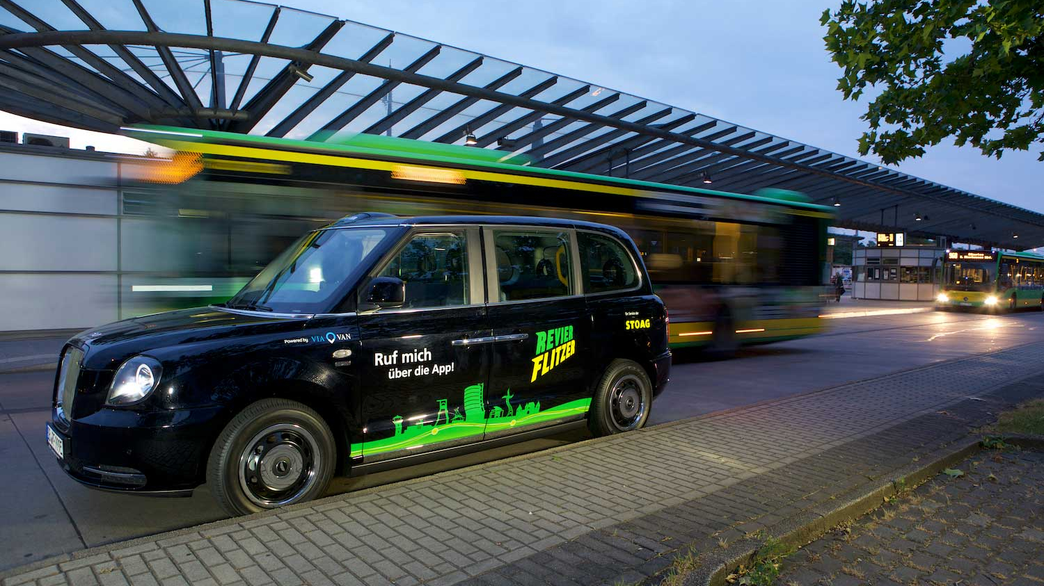 Ein kleines schwarzes Auto mit dem Aufkleber Revierflitzer steht an einer Haltestelle, im Hintergrund fahren gelb-grüne Busse.