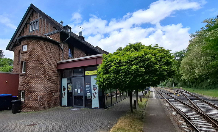 Historisches Backsteingebäude am Bahnhof Heimbach mit Bahnsteig und Gleisen, Baumreihe und blauer Himmel, Eingang zur Regionalbahn