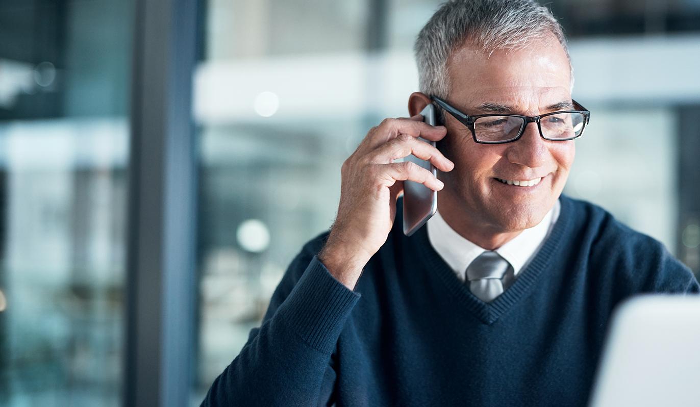 Header FAQ Ein Man sitzt an einem PC und telefoniert gleichzeitig mit einem Smartphone.