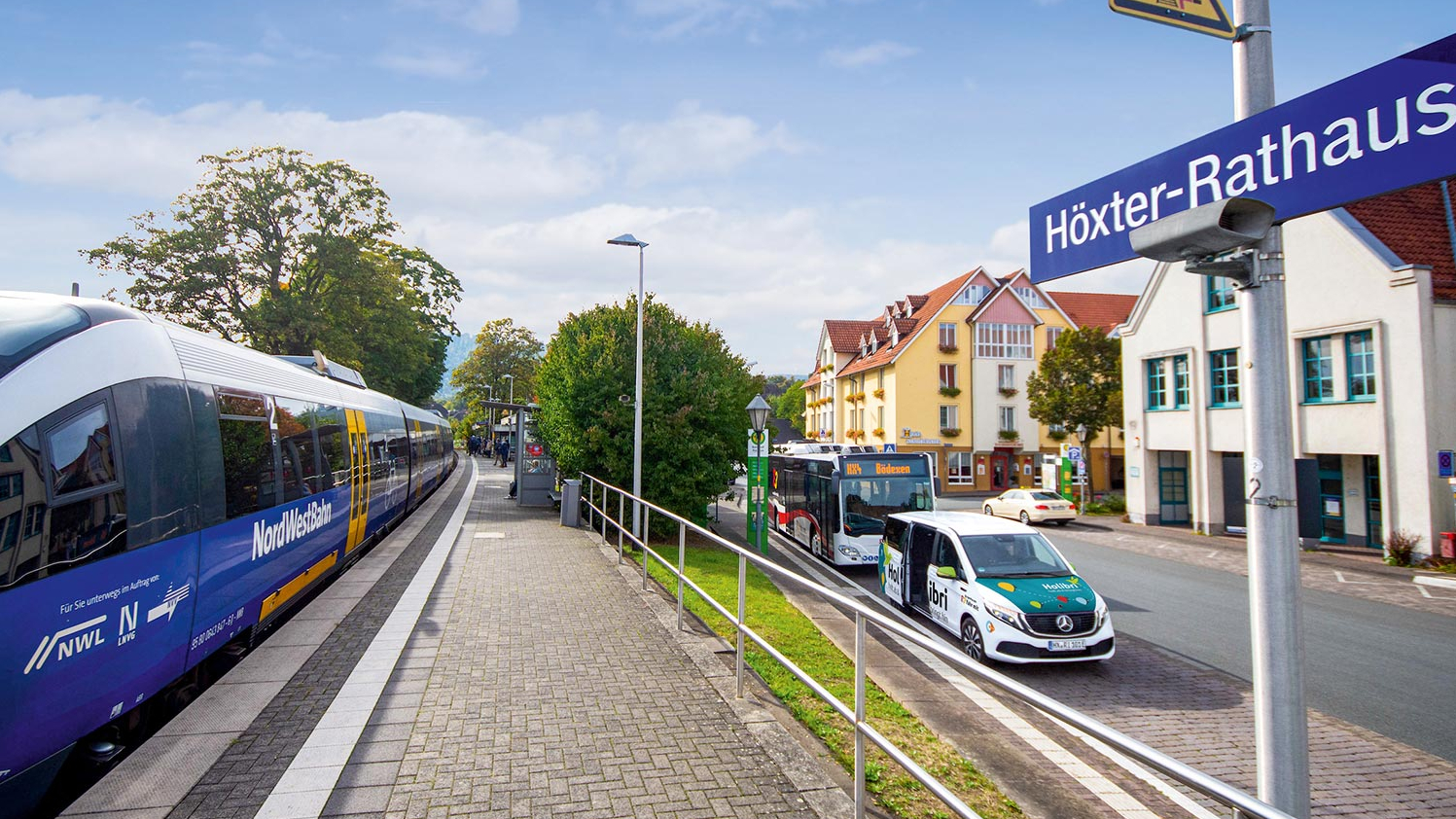 Ein Zug steht an einem Bahngleis, auf der Straße daneben parken ein Holibri-Fahrzeug und ein Linienbus.