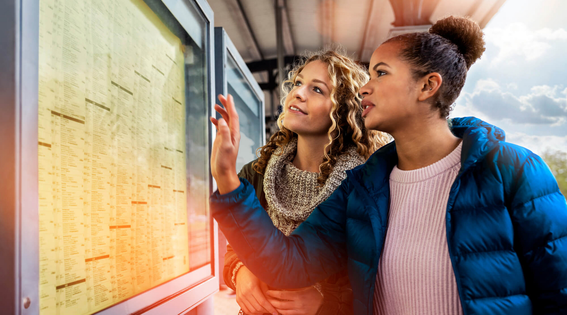 Zwei junge Frauen stehen an einem Bahnsteig und betrachten den Abfahrtsplan.