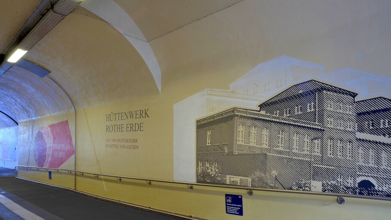 Die Bahnstation am Bahnhof Rothe Erde in Aachen, die mit Bildern des Hüttenwerks Rothe Erde verziert ist.