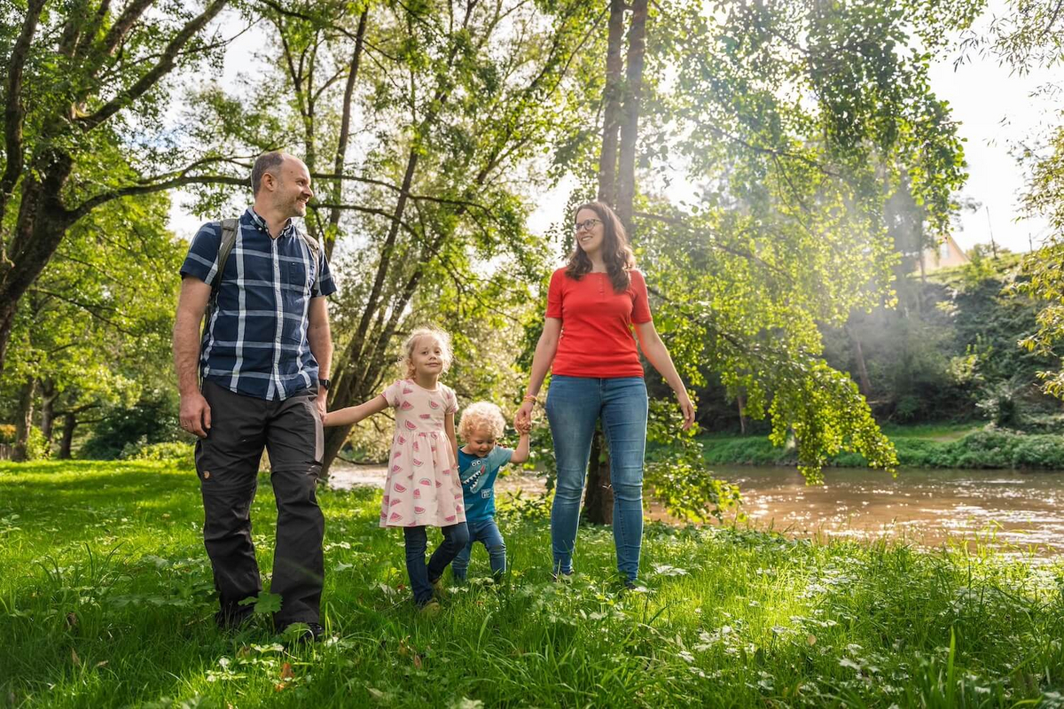 Ein Mann in blauem Hemd und eine brünette Frau mit rotem Shirt halten zwei kleine blonde Kinder an den Händen. Sie laufen über eine Wiese. Die Sonne scheint.