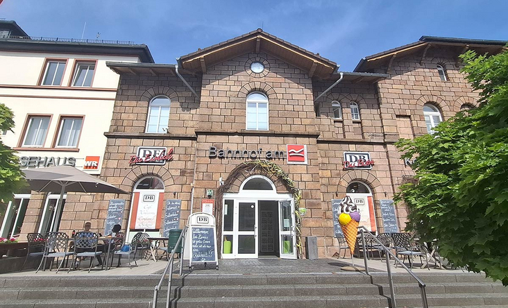 Historisches Bahnhofsgebäude Lennestadt‑Altenhundem mit Außengastronomie und DB‑Service, sonnige Treppenanlage, Willkommensschilder am Eingang