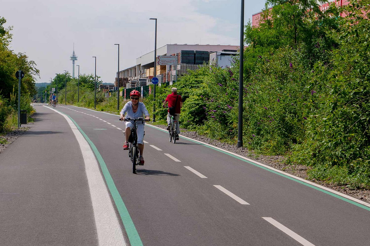 Eine Frau mit rotem Helm fährt an einem sonnigen Tag über einen asphaltierten Radweg durchs Grüne.