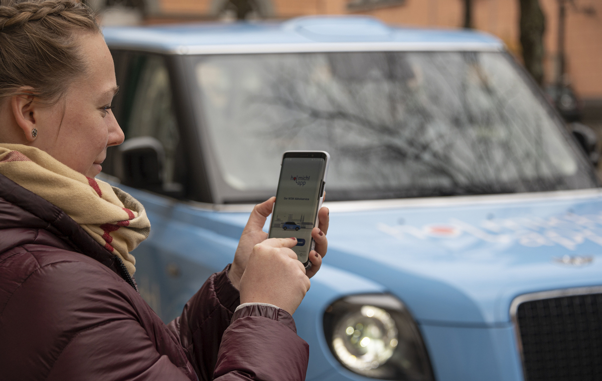 Eine Frau mit einem Smartphone in der Hand steht vor einem kleinen blauen Auto. Auf dem Handybildschirm ist die hol mich! app zu sehen.