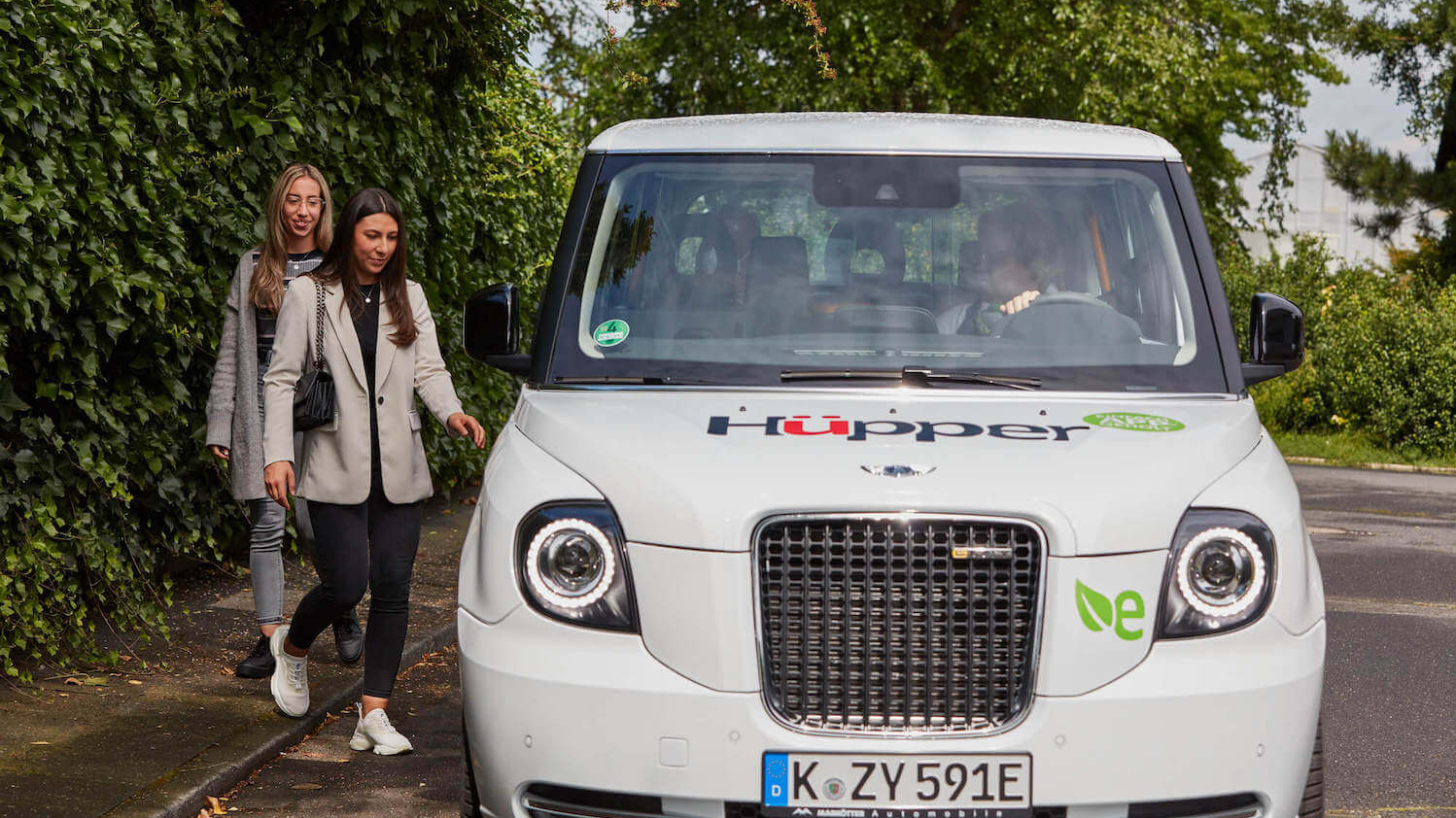 Ein weißes Fahrzeug in Frontansicht. Daneben stehen zwei lächelnde junge Frauen, die eine greift nach dem Türgriff des Autos.