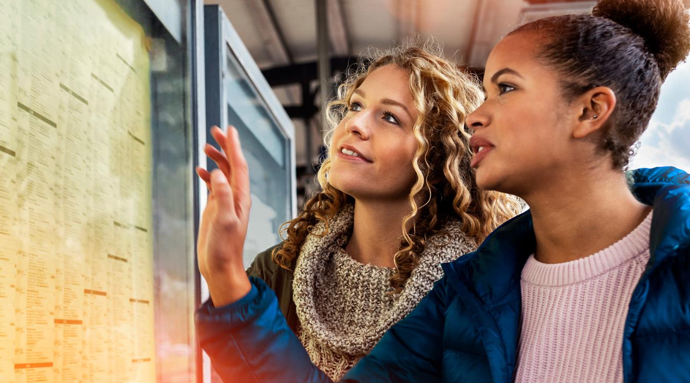 Header Fahrplanauskunft Zwei junge Frauen stehen an einem Bahnsteig und betrachten den Abfahrtsplan.