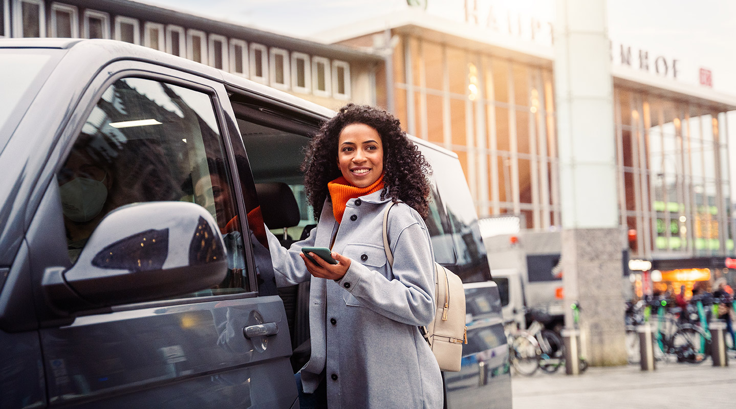 Eine junge Frau steigt an einem Bahnhof lächelnd in ein On-Demand-Fahrzeug.