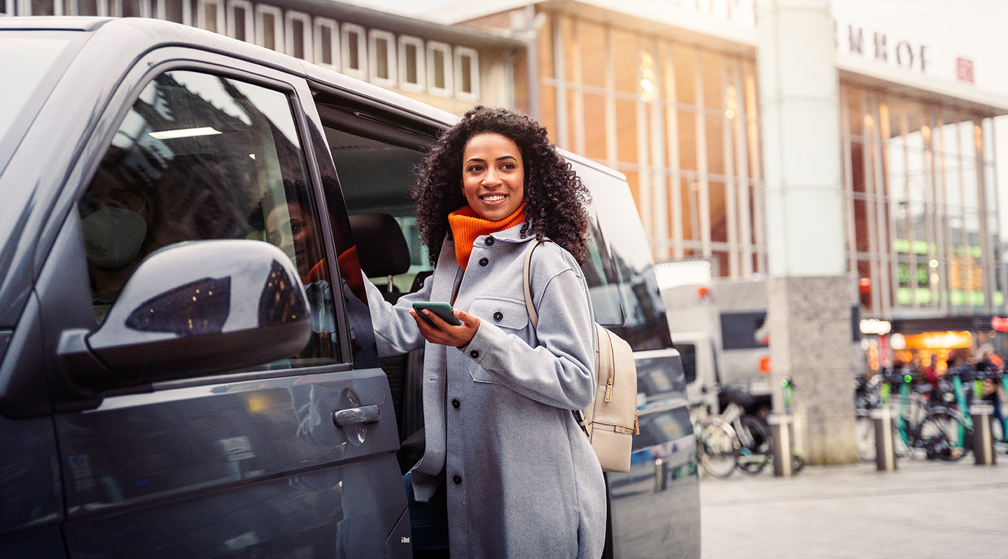 Eine junge Frau steigt an einem Bahnhof lächelnd in ein On-Demand-Fahrzeug.