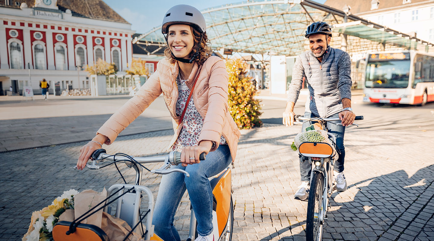 Eine Frau und ein Mann mittleren Alters fahren auf Leihrädern über einen Platz vor dem Hammer Hauptbahnhof.