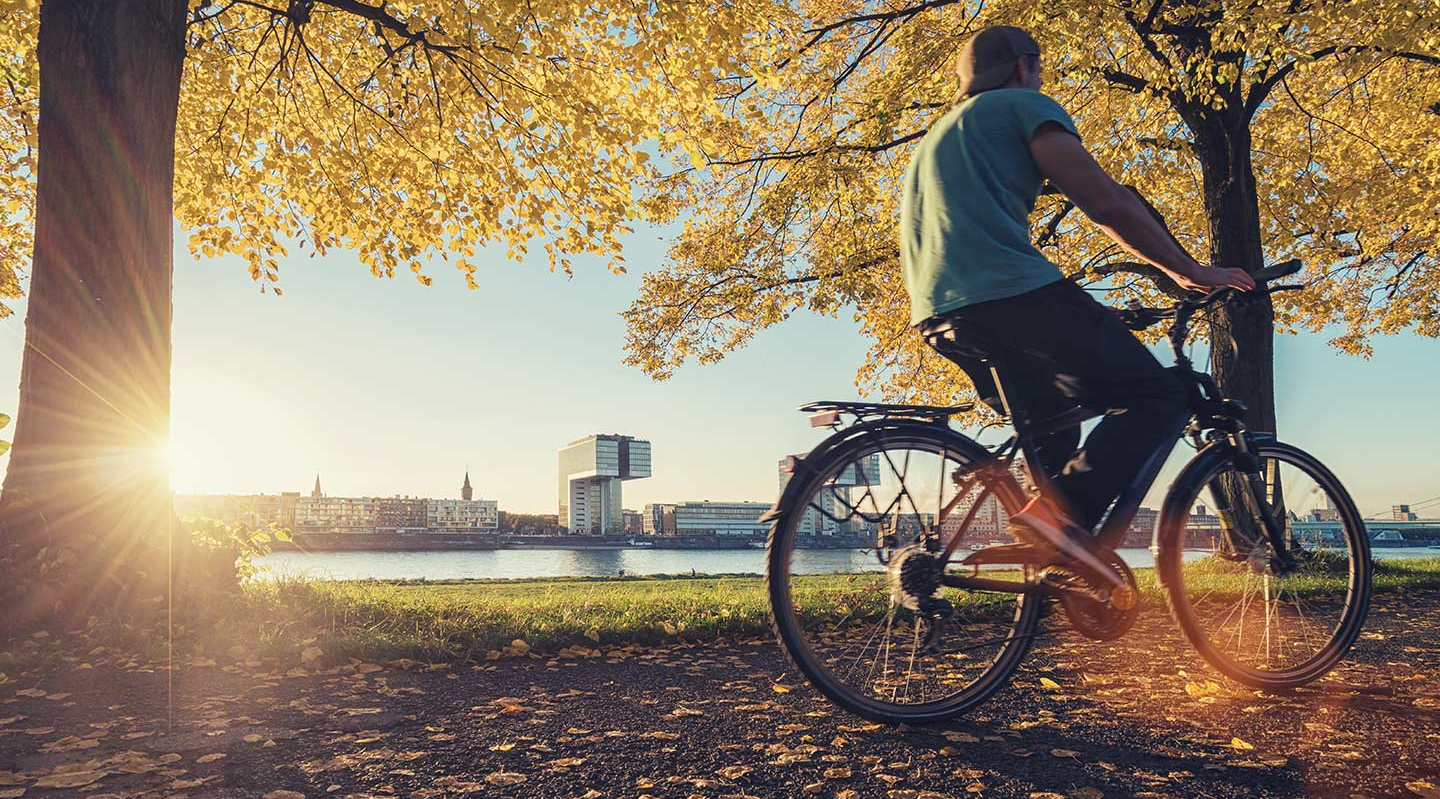 Ein junger Mann fährt im Herbst auf einem Fahrrad am Rhein vor den Kölner Kranhäusern vorbei.
