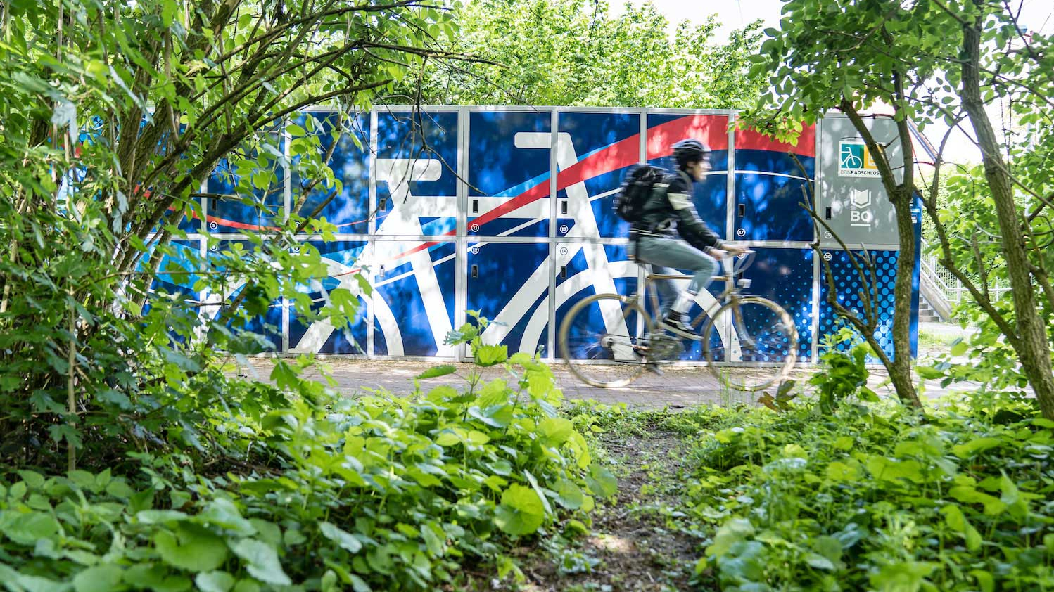 Ein Mann fährt auf einem Fahrrad an einer blauen Radbox vorbei.