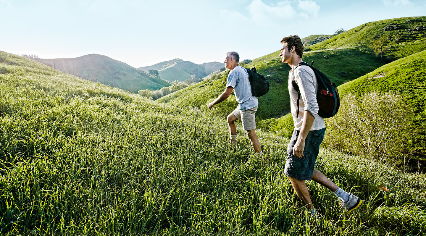 Header Wunderbar wanderbar 2020 Ein jüngerer und ein älterer Mann wandern im Sommer von rechts nach links über eine hügelige Graslandschaft.