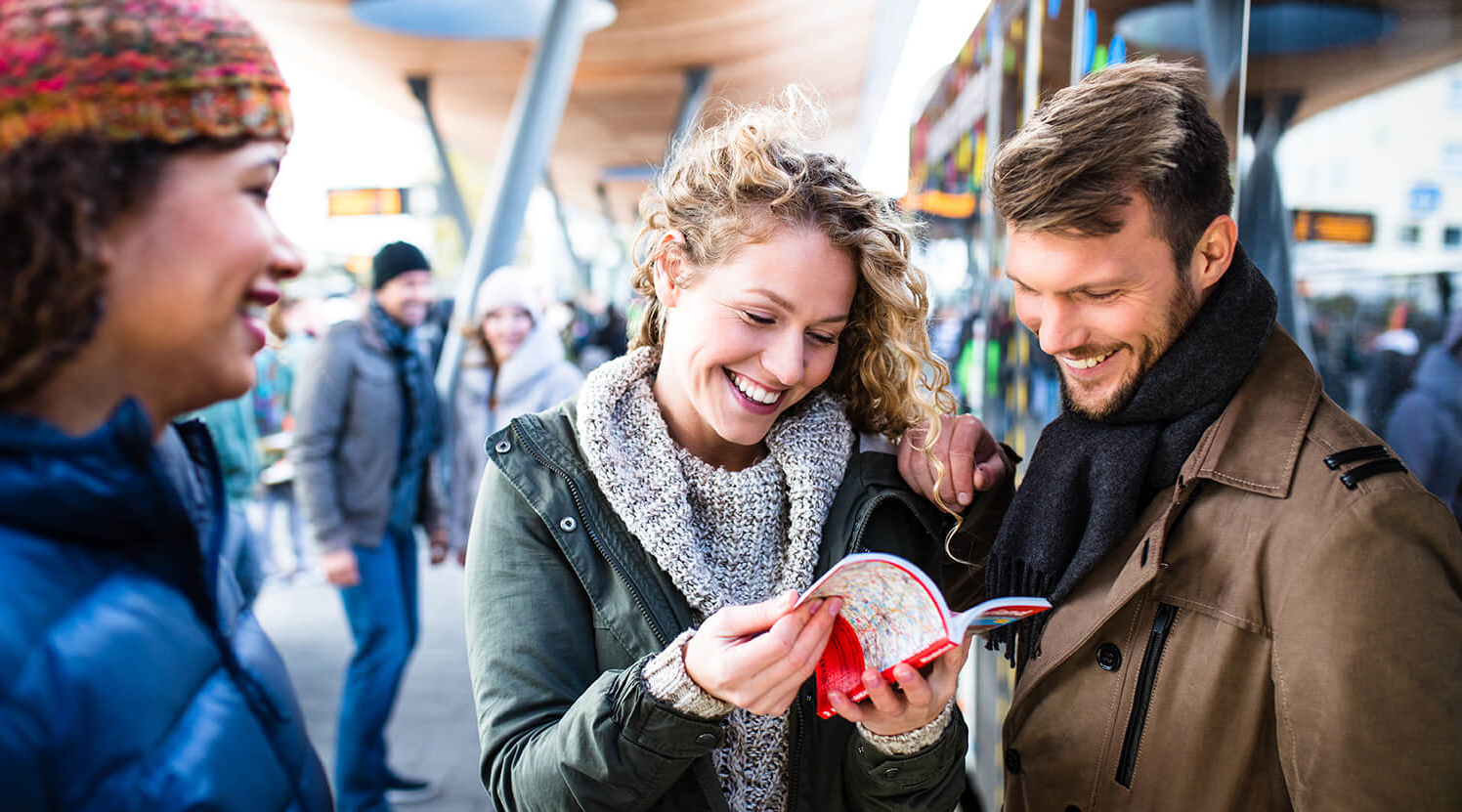 Eine Gruppe von Personen steht lachend an einem Busbahnhof und blättert in einem Ticketflyer.