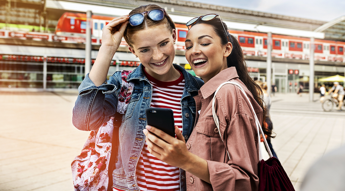 Zwei Schülerinnen in Sommerkleidung stehen vor einem Bahnhof und schauen fröhlich auf ein Handy.