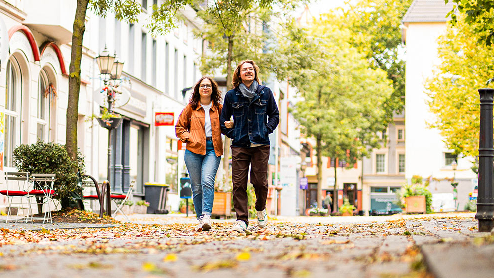 Zu sehen sind zwei Menschen, welche zusammen durch eine herbstliche Stadt-Szenerie laufen. 