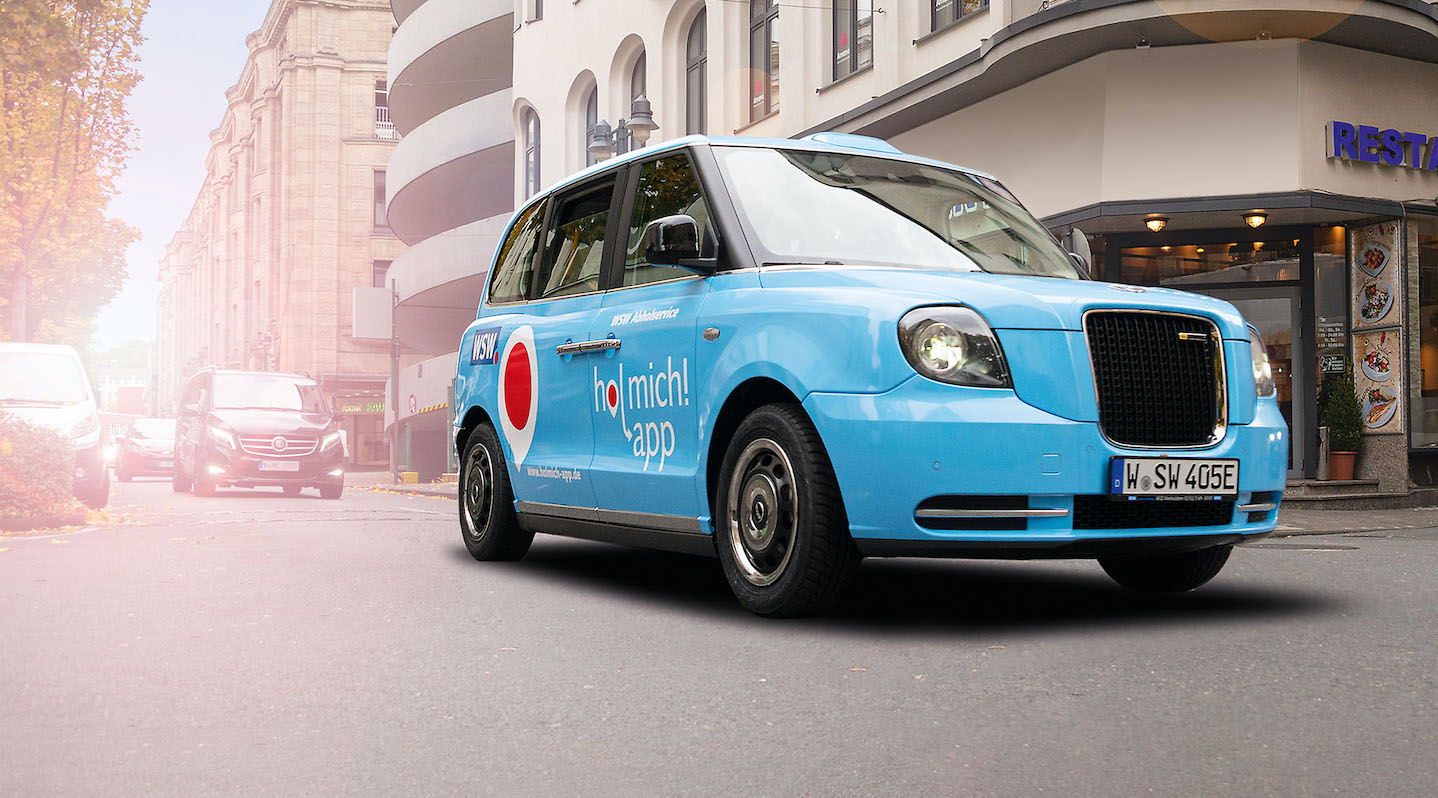 Ein kleines blaues Auto mit dem Schriftzug "hol mich! app" auf der Seitentür fährt über eine Straße.