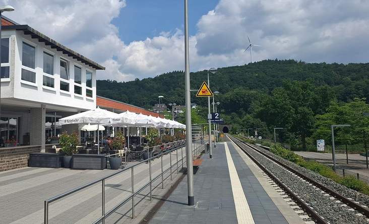 Bahnhof Sondern mit Außenterrasse am Bahnsteig, Blick auf Gleis 2 und bewaldete Hügel, sommerlicher Himmel mit Wolken