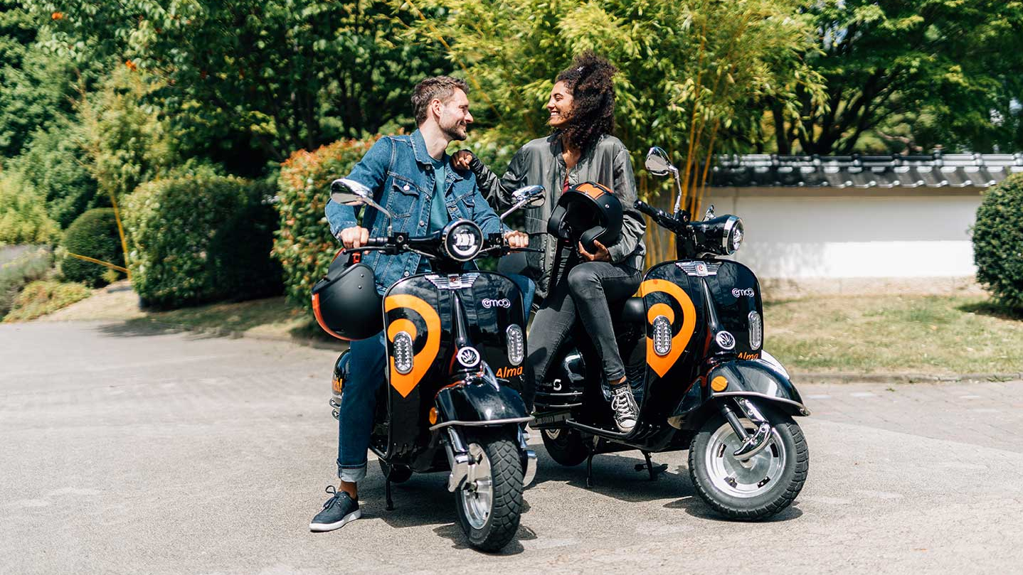 Ein junger Mann und eine junge Frau sitzen auf zwei orange-schwarzen E-Rollern mit der Aufschrift Alma.