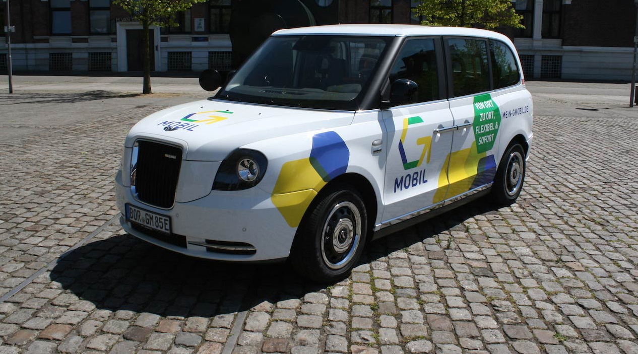 Ein kleines weißes Auto steht auf einem Kopfsteinplatz. Auf das Auto wurden gelbe und blaue Sechsecke lackiert, außerdem ist das Logo von G-Mobil zu sehen.