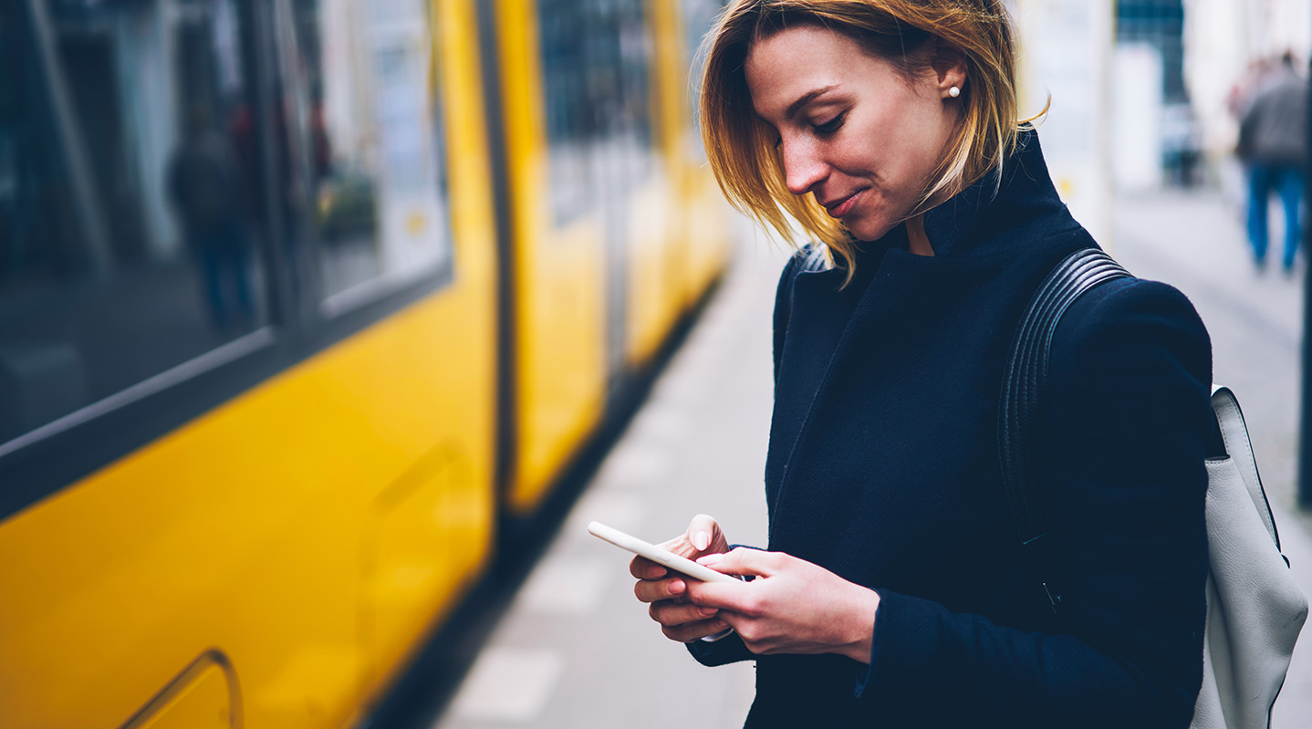 Eine junge Frau steht an einer Straßenbahn und schaut auf ihr Handy.