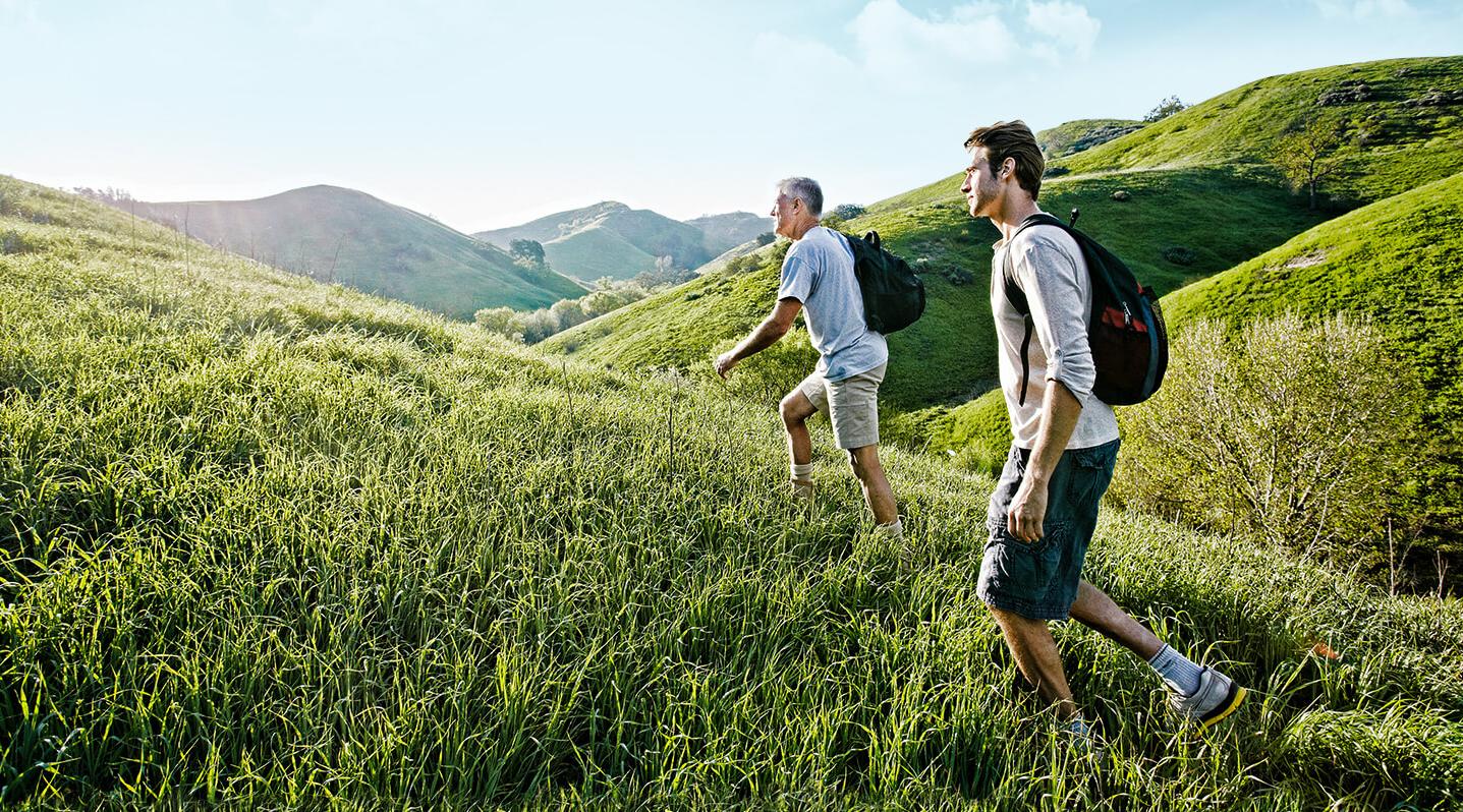 Header Wunderbar wanderbar 2020 Ein jüngerer und ein älterer Mann wandern im Sommer von rechts nach links über eine hügelige Graslandschaft.