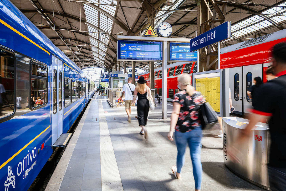 Sommerlich gekleidete Personen laufen an einem überdachten Bahnsteig an einem blauen Zug vorbei.